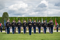 Members of the Army are standing at attention on a green lawn in front of a dark green hedge. Their uniform is top is black with lighter blue pants and black hats. The first member has a sword.