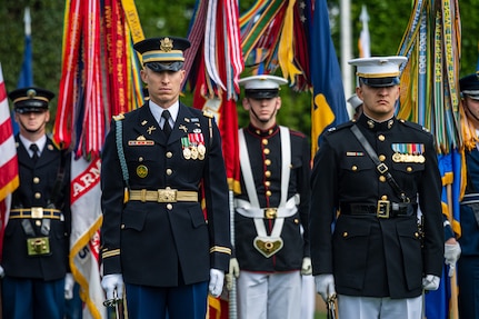 Service members in various types of military dress uniforms are standing at attention on a green lawn in front of a dark green hedge with flags and streamers behind them.