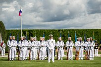 Members of the Navy are standing at parade rest on a green lawn in front of a dark green hedge with the US and Philippines flags flying behind them. Their uniform is white with white hats. Each member has a rifle in front of them, except the first member who has a sword.