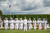 Members of the Navy are standing at parade rest on a green lawn in front of a dark green hedge with the US and Philippines flags flying behind them. Their uniform is white with white hats. Each member has a rifle in front of them, except the first member who has a sword.