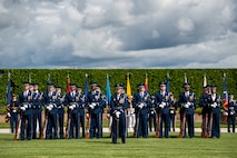 Members of the Air Force are standing at parade rest on a green lawn in front of a dark green hedge with the US and Philippines flags flying behind them. Their uniform is dark blue with dark blue hats. Each member has a rifle in front of them, except the first member who has a sword.