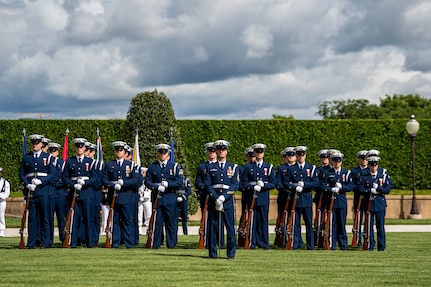 Coast Guard members are standing at parade rest on a green lawn in front of a dark green hedge with the US and Philippines flags flying behind them. Their uniform is dark blue with white hats. Each member has a rifle in front of them, except the first member who has a sword.