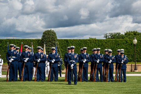 Coast Guard members are standing at parade rest on a green lawn in front of a dark green hedge with the US and Philippines flags flying behind them. Their uniform is dark blue with white hats. Each member has a rifle in front of them, except the first member who has a sword.