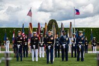 Service members in various types of military dress uniforms are standing at parade rest on a green lawn in front of a dark green hedge with the US and Philippines flags flying behind them.