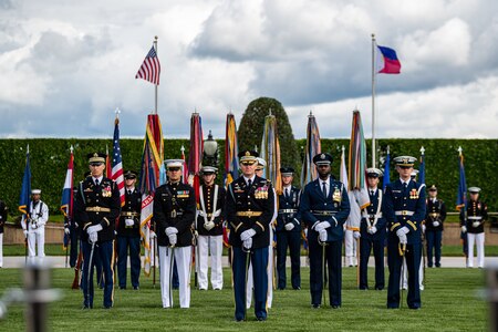 Service members in various types of military dress uniforms are standing at parade rest on a green lawn in front of a dark green hedge with the US and Philippines flags flying behind them.