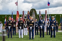 Service members dressed in various types of military dress uniforms are standing at parade rest on a green lawn with a green hedge in the background and the US flag and the Philippines flag flying behind.