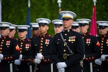 Dozens of Marines dressed in their dress uniforms with black tops and white hats are standing at parade rest in front of a dark green hedge.