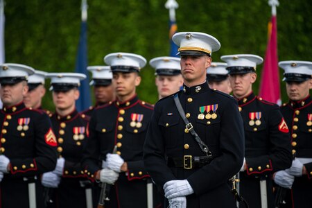 Dozens of Marines dressed in their dress uniforms with black tops and white hats are standing at parade rest in front of a dark green hedge.