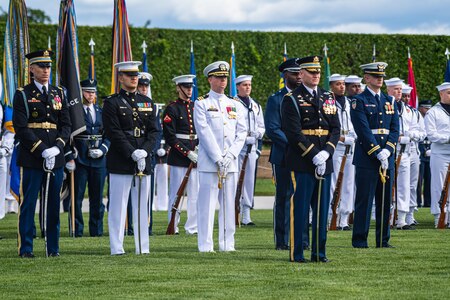 Members in various types of military dress uniforms stand at parade rest in front of a large green hedge on a green lawn.