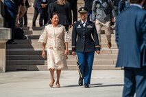 Two women smiling as they walk together toward the camera, away from steps where other peoeple are congregating. The woman on the left is in a gold-colored dress and on the right is wearing an Army dress uniform.