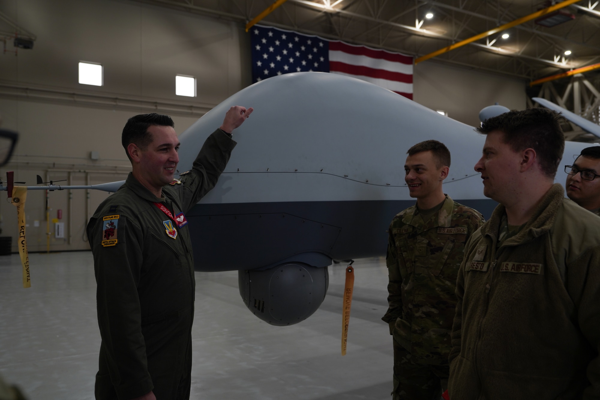 A man in a flight suit with his hand on a remotely piloted aircraft talks to men in military uniforms.