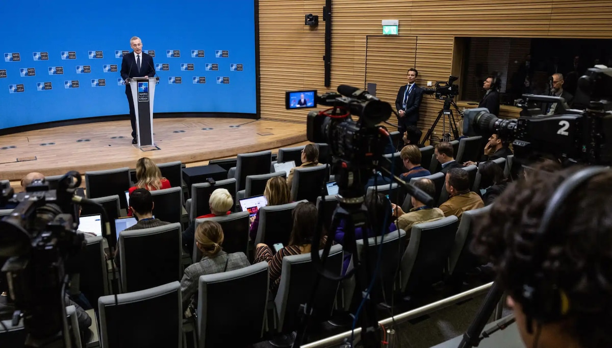 A man in business attire stands behind a lectern on a stage and addresses an audience. The backdrop indicates that he is at NATO.