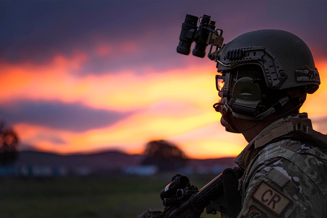 An airman wearing a helmet looks into the distance on a flight line, with a pinkish purplish sky as backdrop.