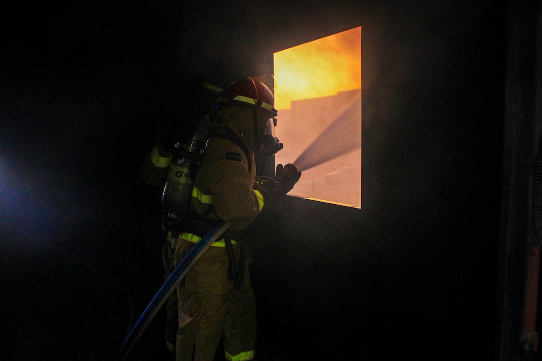 A firefighter in a dark room sprays water from a hose through a rectangular opening revealing a fire.