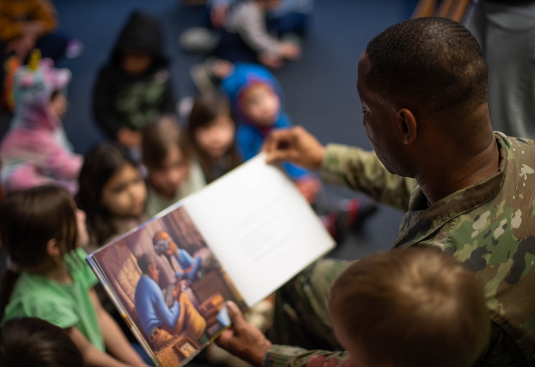 An airman reads a book to children seated on the floor.