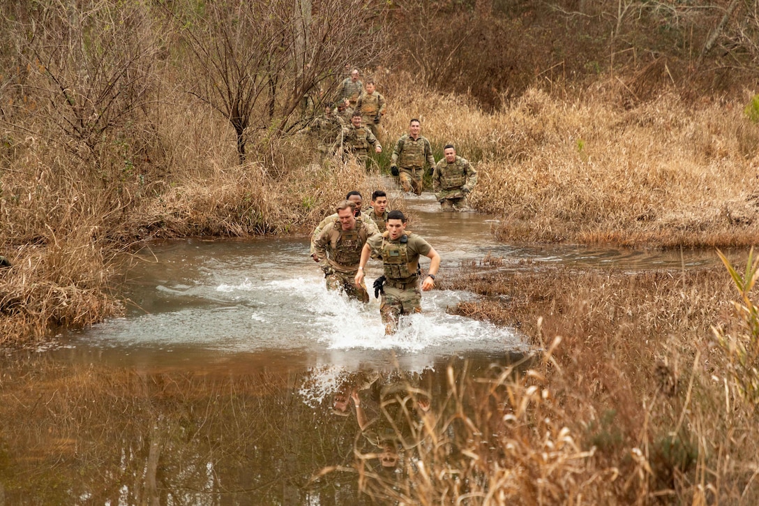 A group of soldiers run through a small river surrounded by brush.