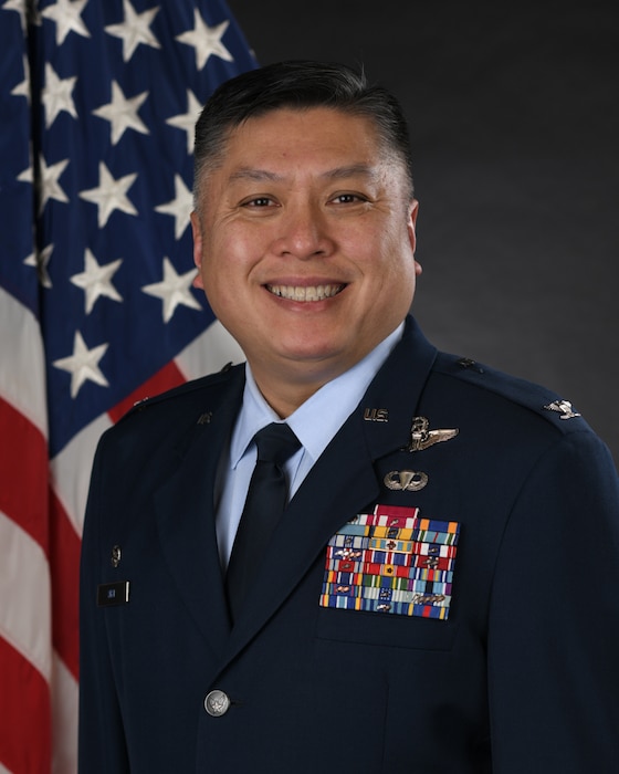 A man, visible from the chest upwards, in a U.S. Air Force service dress uniform. He is smiling and his uniform contains many different ribbons and metallic objects, including his name tag that reads "THAI". In the background, an American flag.
