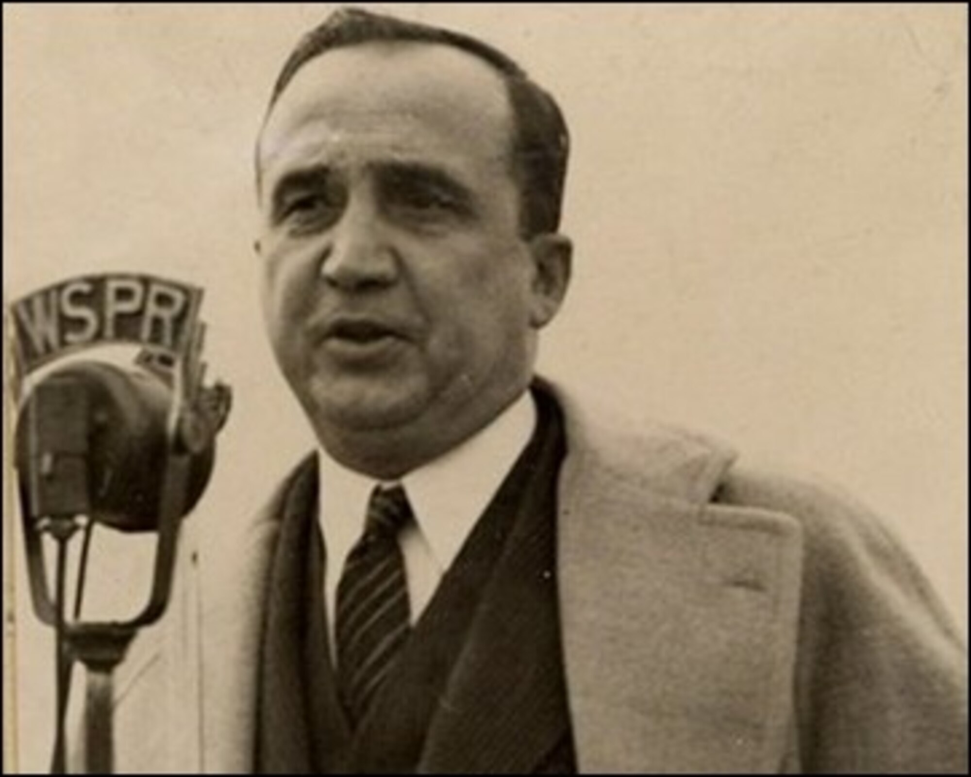 Vintage photo of a man in a suit and overcoat speaking into a public address microphone.