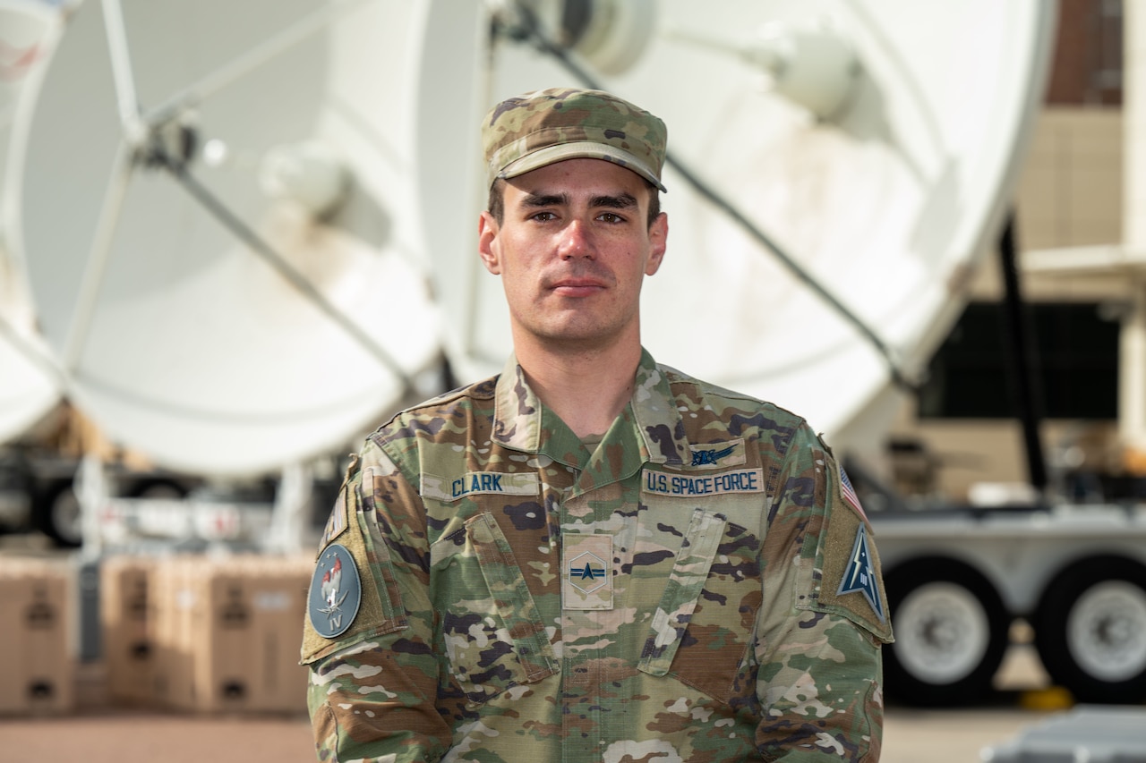 A guardian in camouflage uniform stands outside in front of two white satellite dishes.