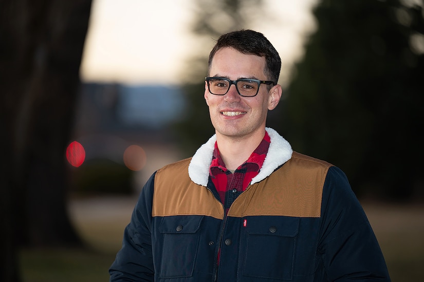 A guardian wearing a red plaid flannel shirt and a jacket smiles for a photo outdoors.