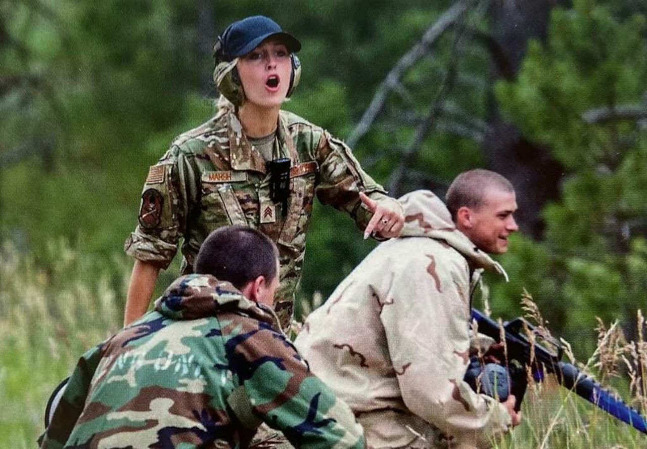 A woman yells orders to two men crouched in a field.