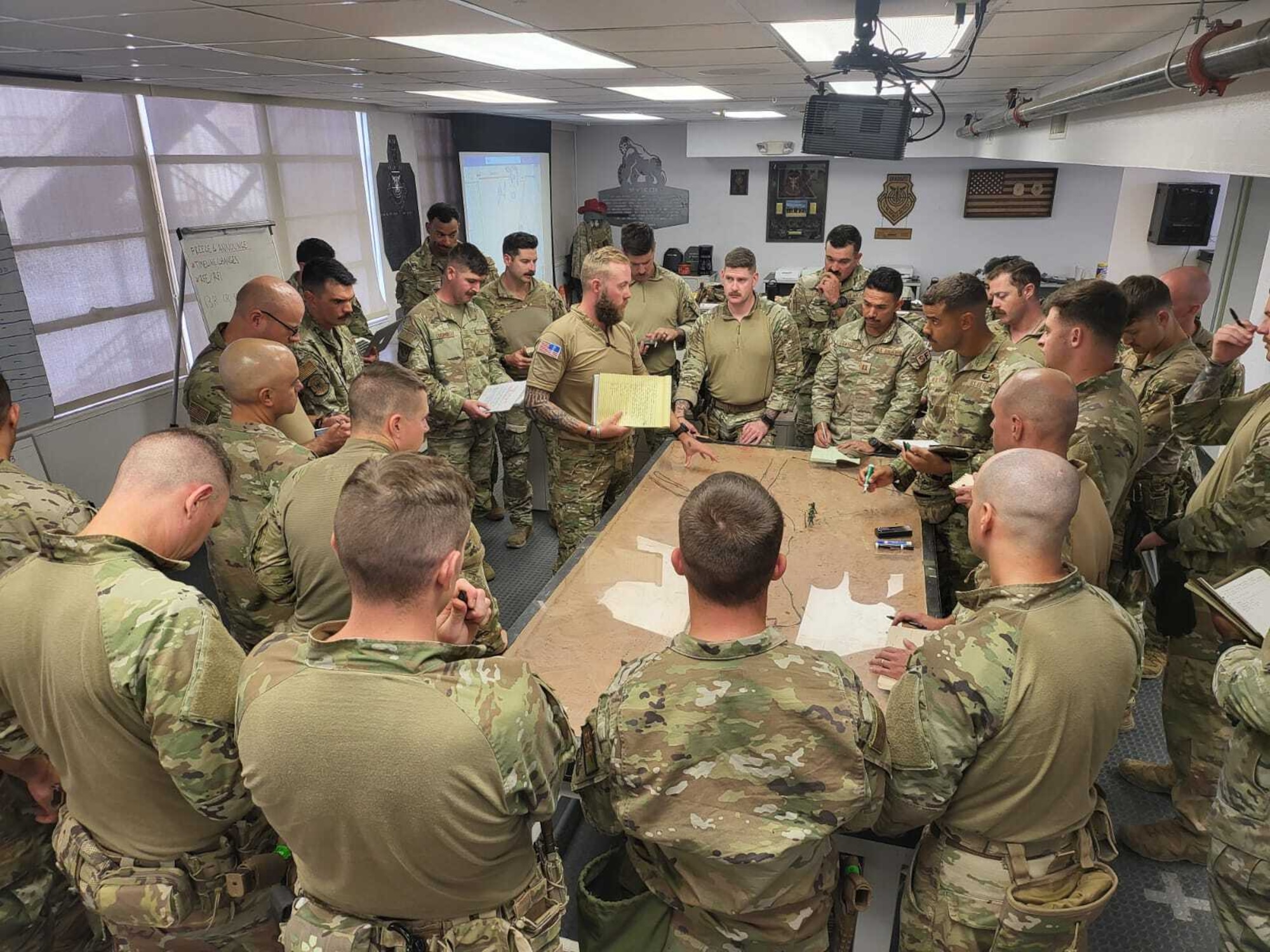 A group of Air Force members in uniform stand in a circle around a map on a table.  One of the persons is telling the others about the map and the upcoming training exercise outdoors.