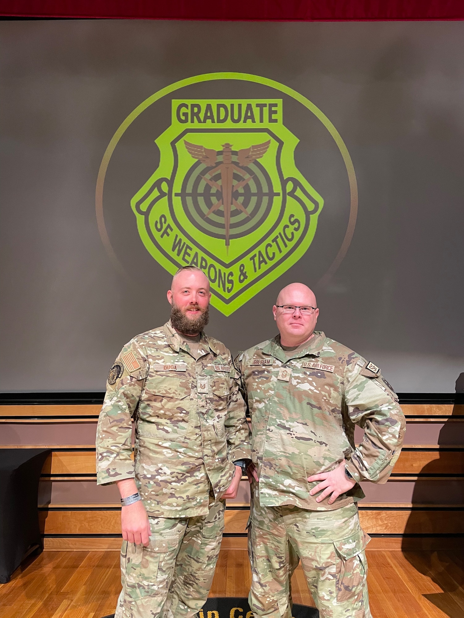 Two Air Force members in uniform stand in front of of a Security Forces Weapons and Tactics emblem.