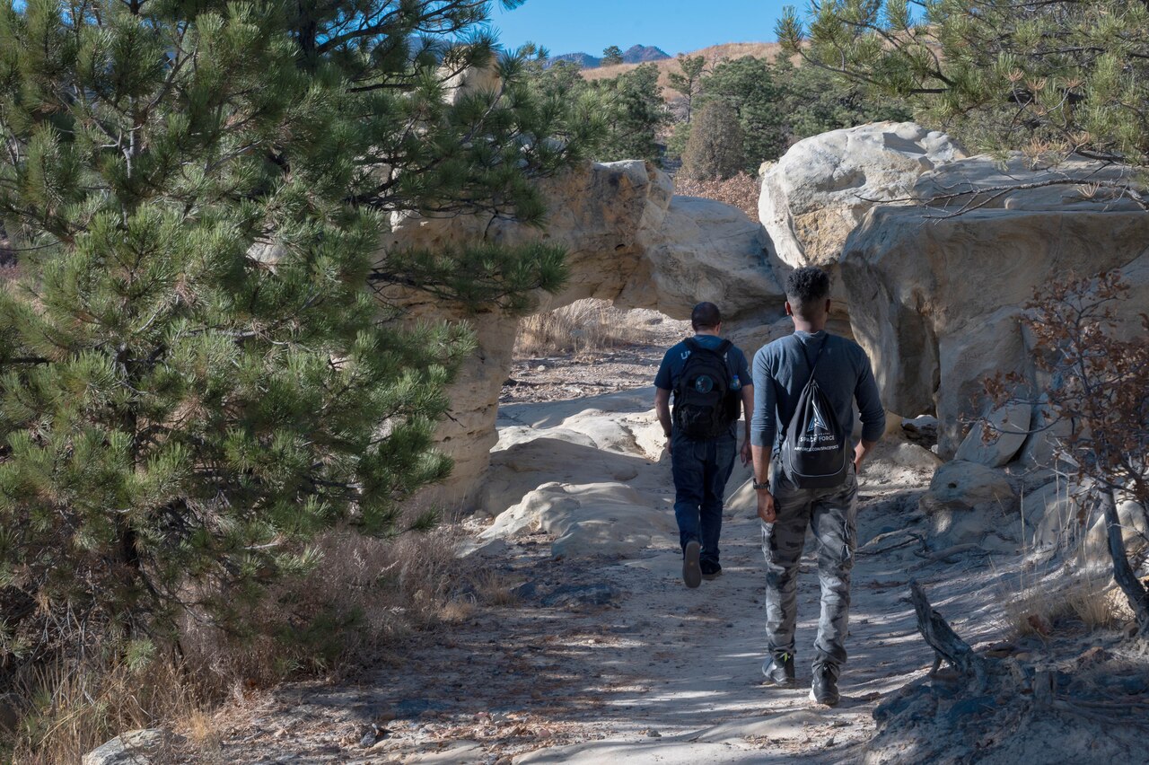 Two guardians in civilian clothes, shown from behind, walk along a dirt trail in the wilderness.