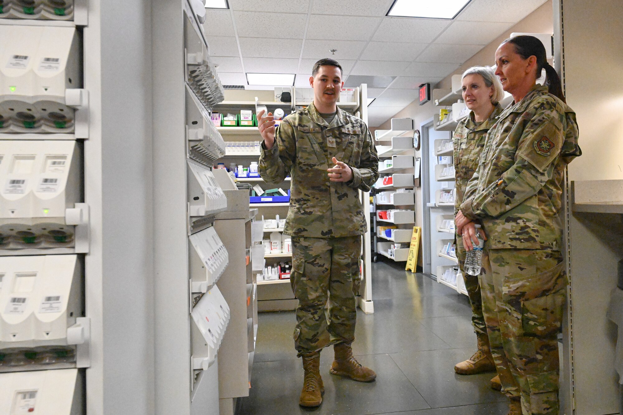 Three Airmen stand in a pharmacy room as one Airman gestures towards shelves and talks.