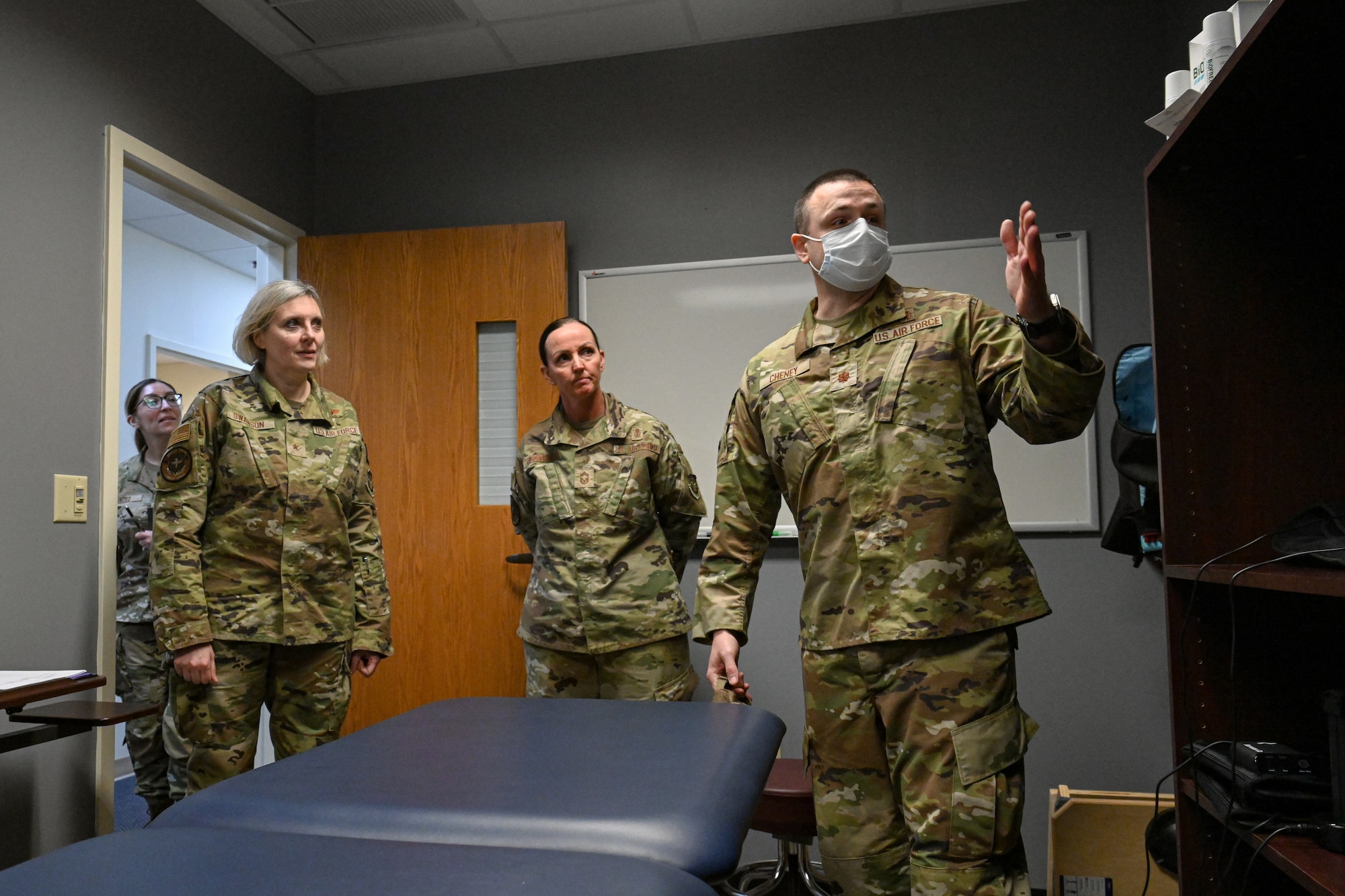 Three Airmen stand in a medical room as one of them points to an object out of frame and talks.