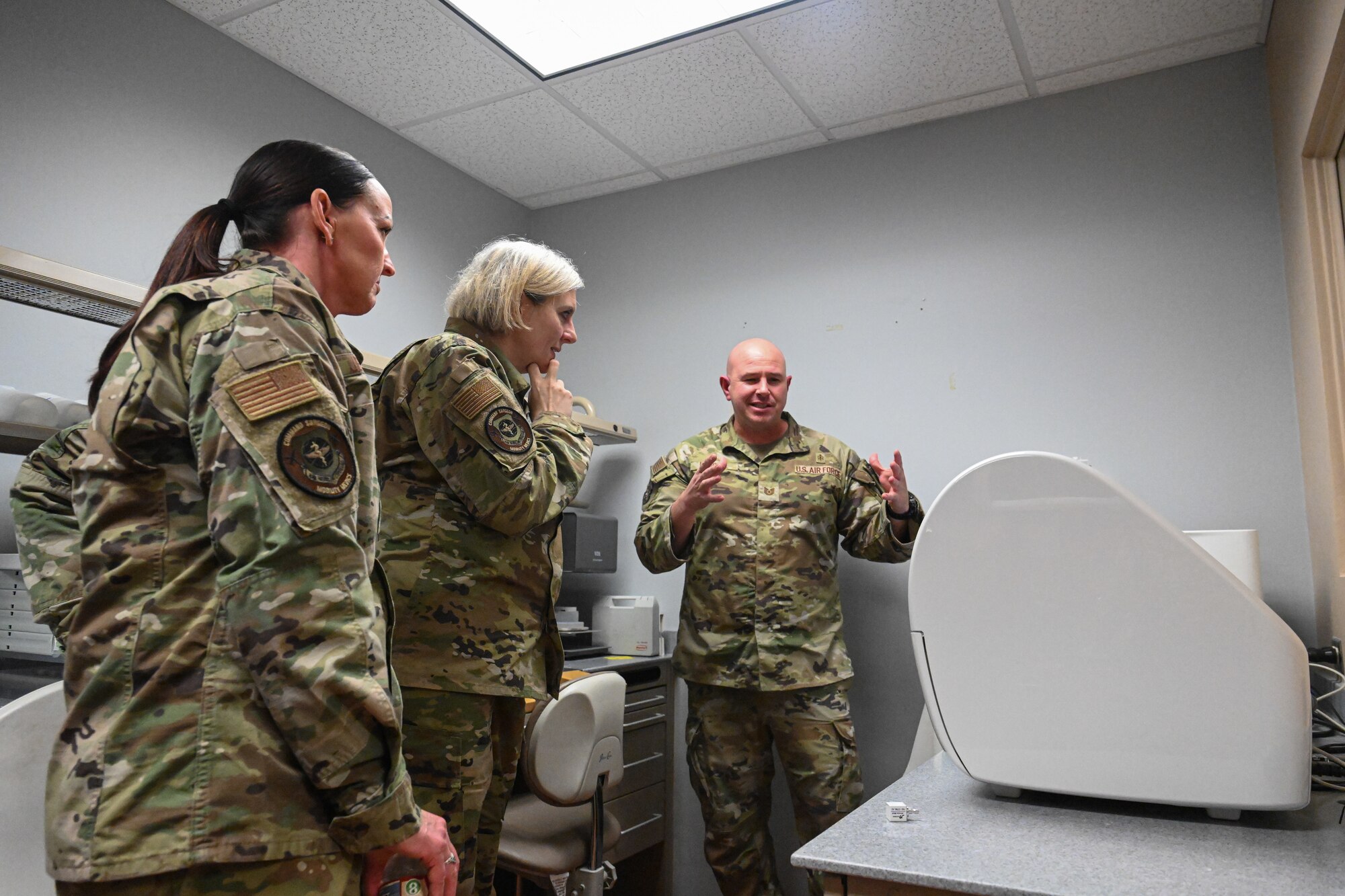 Three Airmen stand in a small medical room and have a discussion.