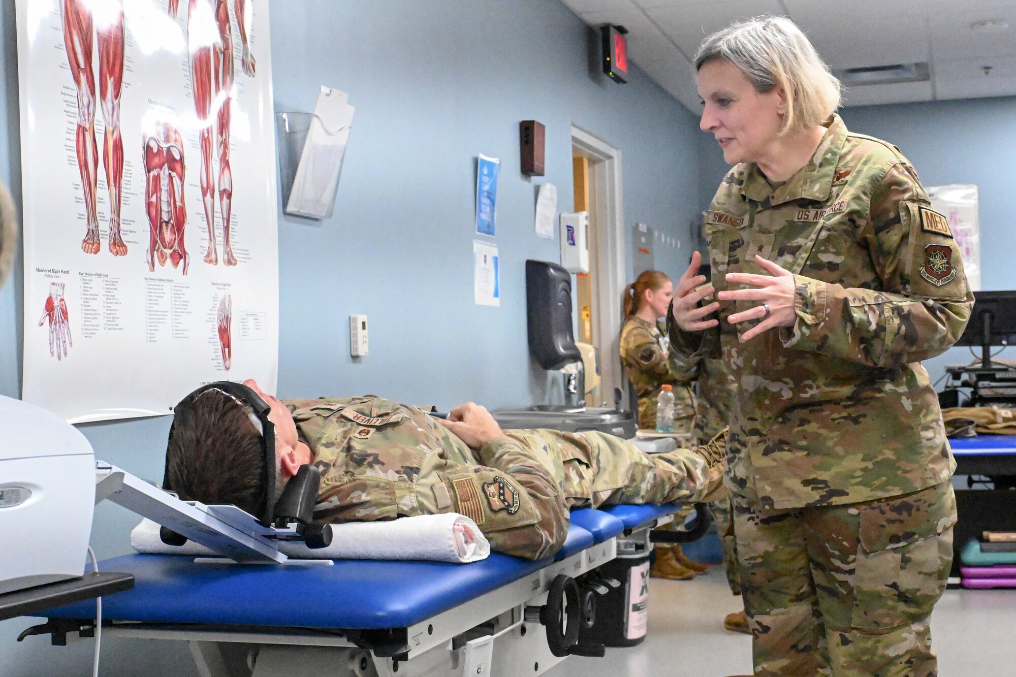 An Airman stands and speaks to another Airman lying on an examination table in a physical therapy room.