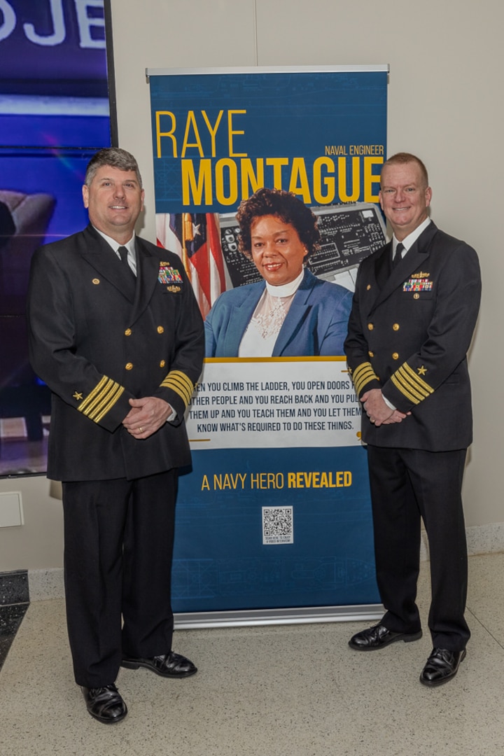 Naval Surface Warfare Center, Carderock Division Commanding Officer Matthew Tardy (left) and former Carderock Commanding Officer Capt. Todd Hutchison stand together next to a banner paying homage to the Navy’s own “hidden figure” Raye Montague.