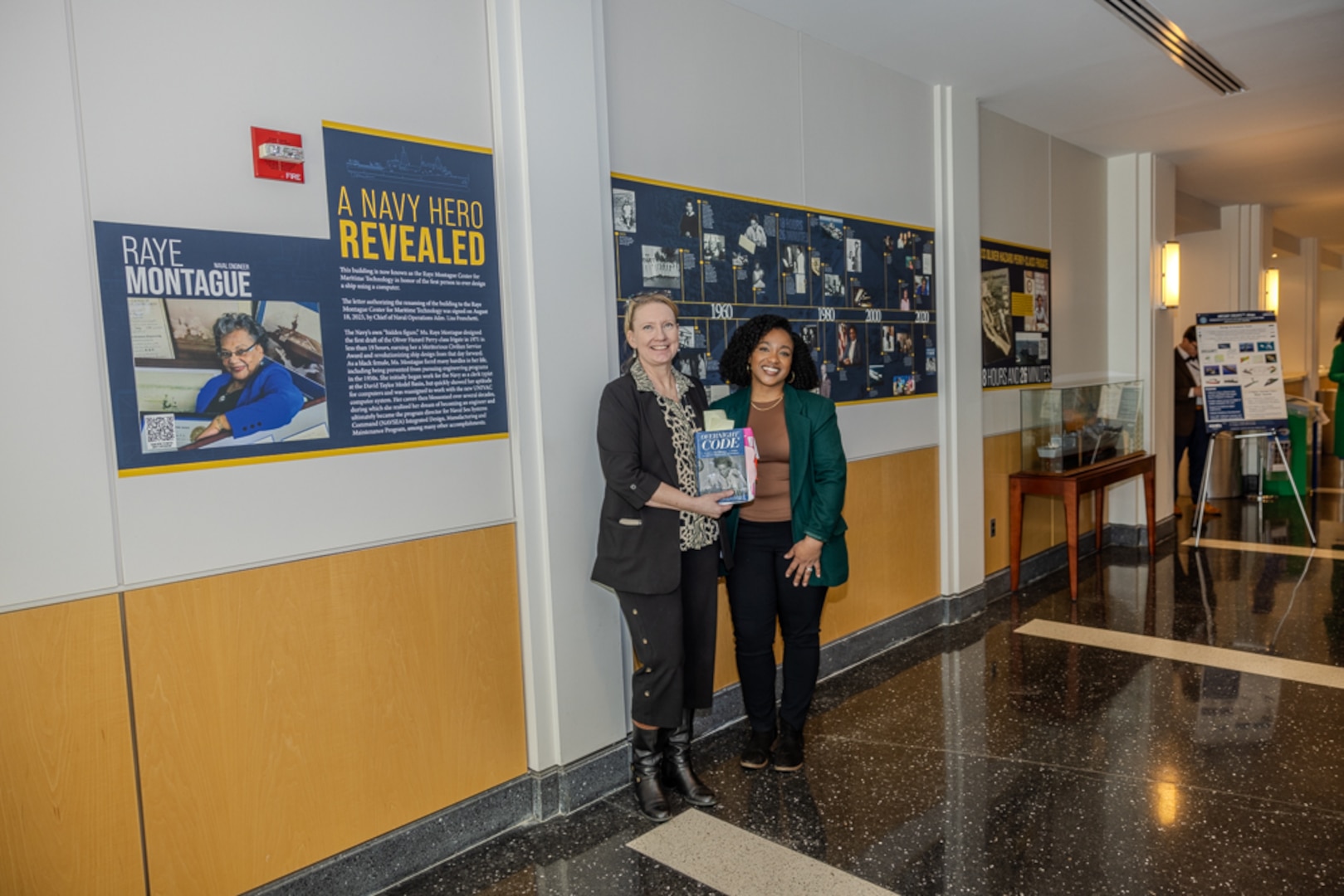 Director for Corporate Communications Kelley Stirling (left) and Multimedia Specialist Chalene “Lena” Simmons (right) stand next to a timeline highlighting the career of Navy “hidden figure” Raye Montague.