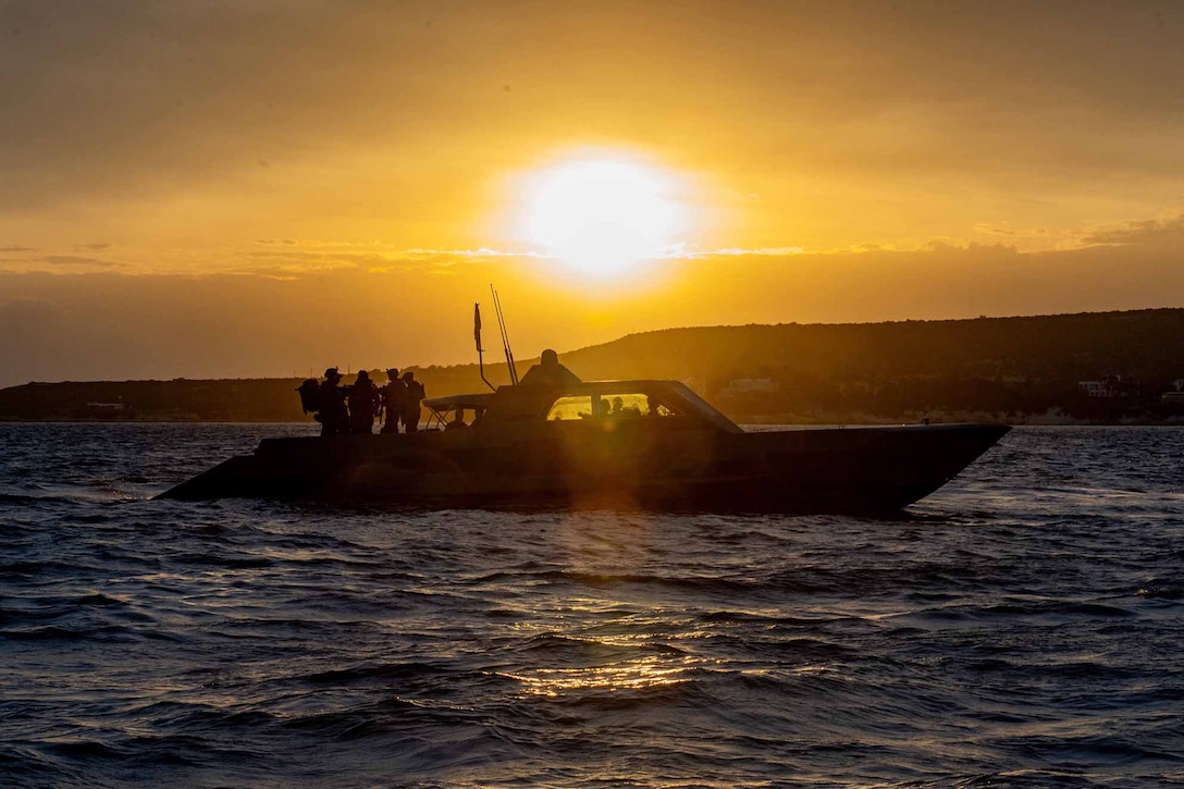 Service members, shown in silhouette, ride in a boat on open water with a low sun.