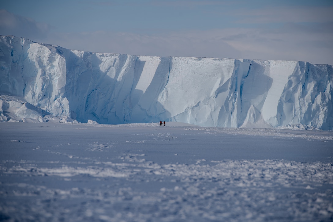Two service members are photographed from a distance walking on snowy ground with a large ice shelf in the background.