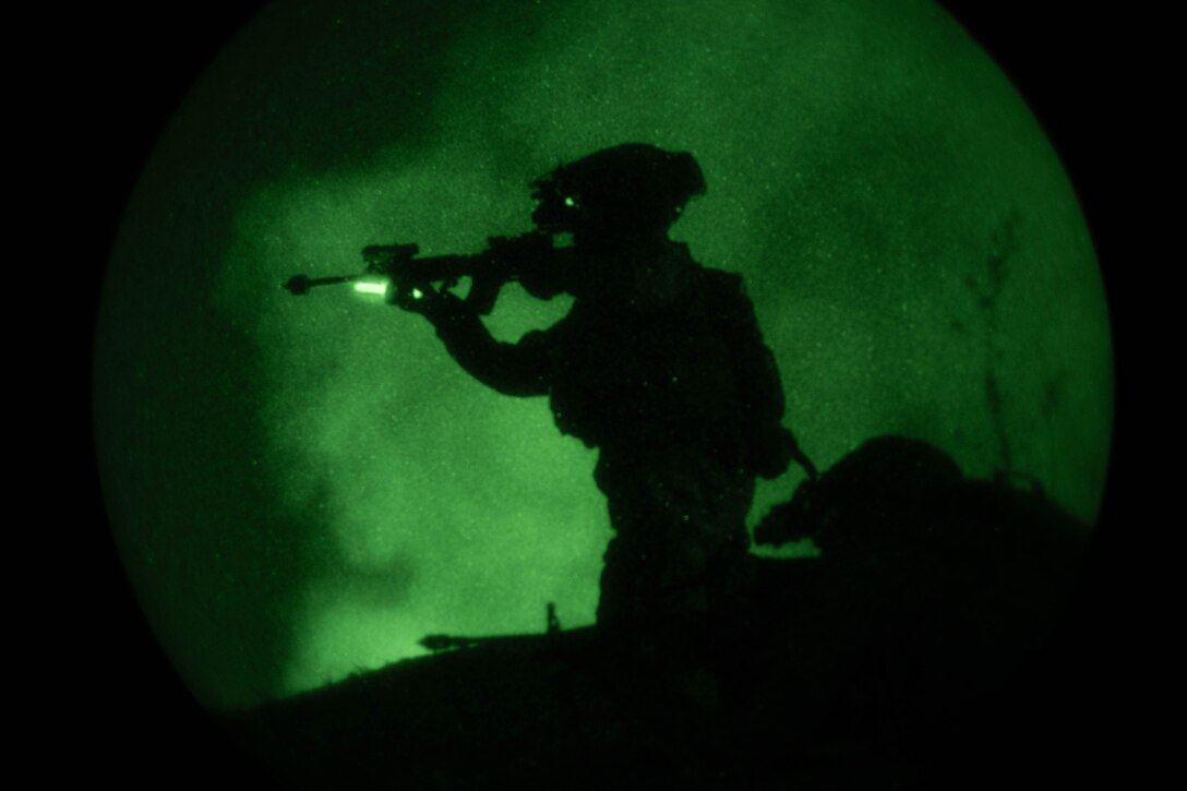 A service member stands while holding a weapon while a second’s helmet is visible closer to the ground. Both are shown in silhouette, illuminated by a green light.