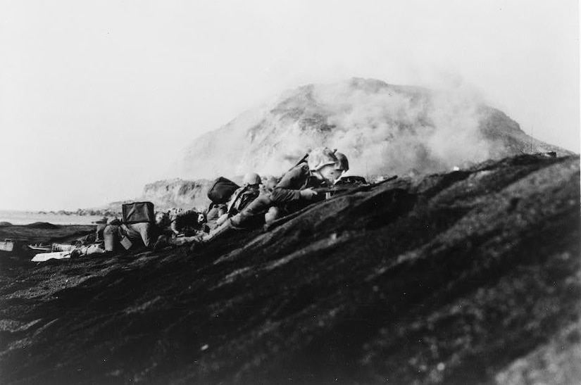 Men lie prone on a beach, aiming their weapons. A mountain looms in the distance.