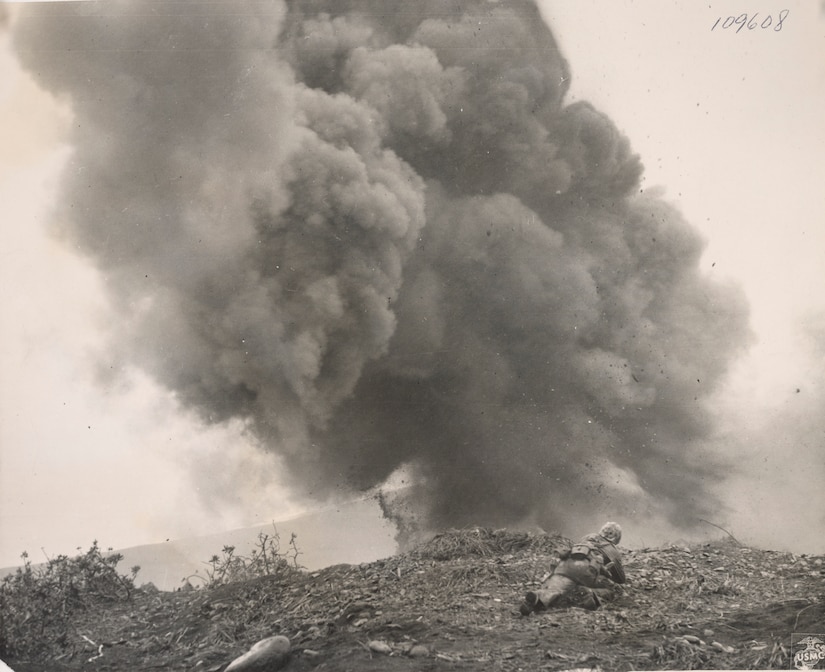A man lies in dirt as a large smoke plume rises into the air in front of him.
