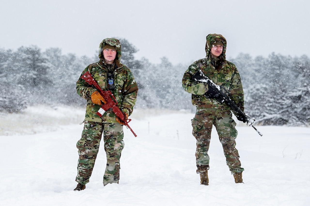Two cadets stand in the snow holding weapons.