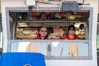 A photo of two men looking out of a boat cabin’s window.