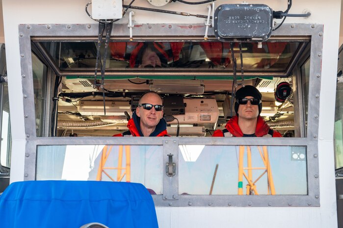 A photo of two men looking out of a boat cabin’s window.