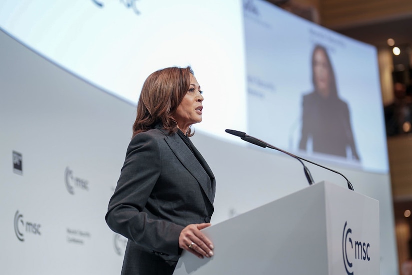 A person in a business suit speaks at a podium with her image projected on a screen in the distance.