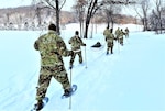 Service members wearing snowshoes and carrying ski poles traverse snow-covered hills.