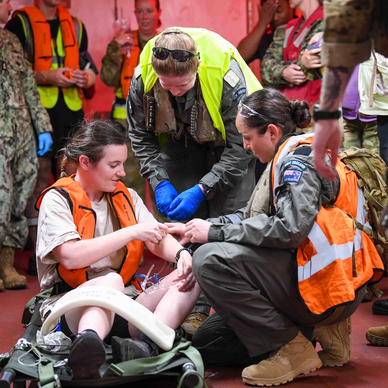 A woman in military uniform administers medical care to another woman. A third woman in military uniform, looks on.  Other military members mull around in the background.