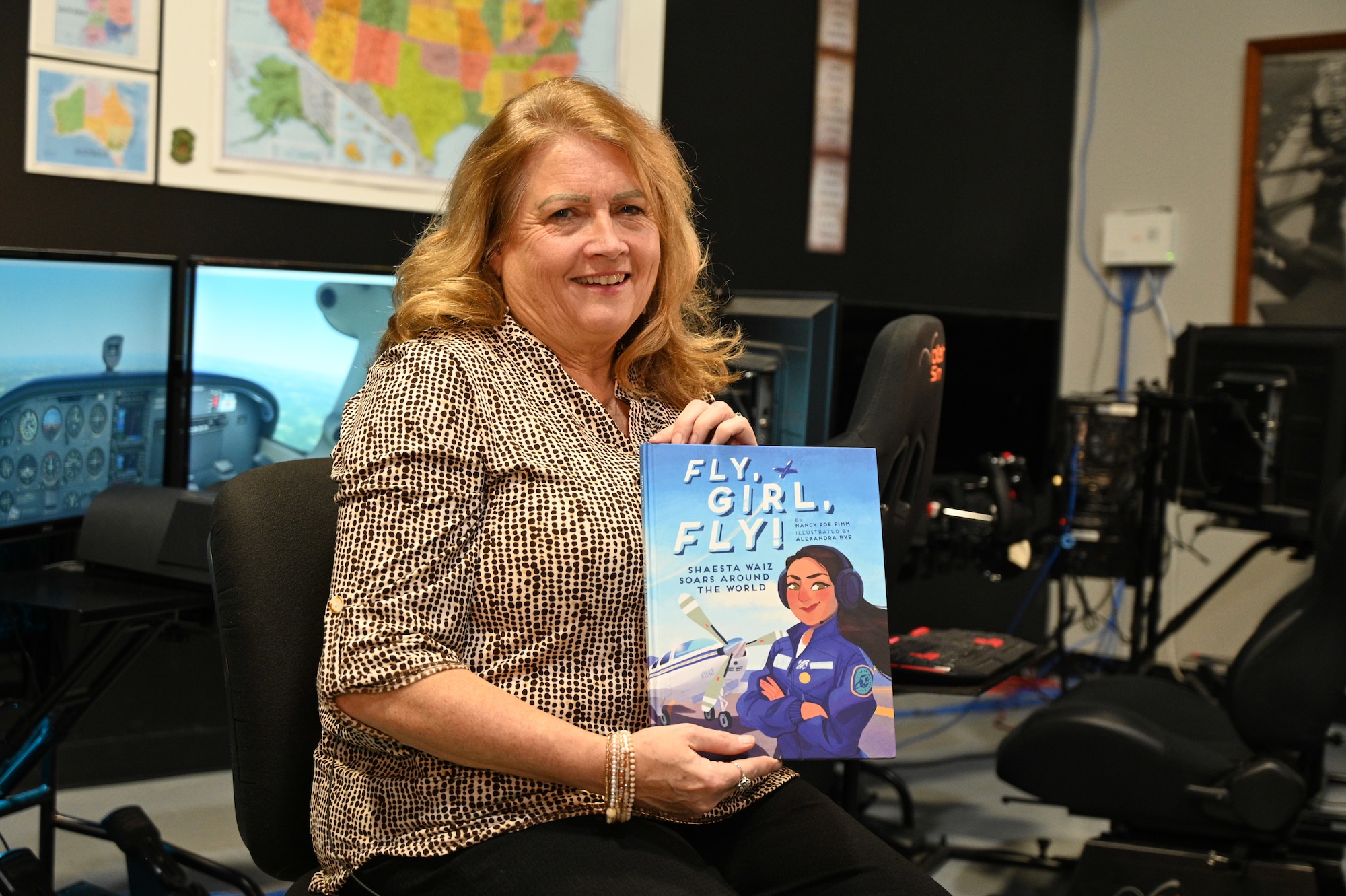 Author Nancy Roe Pimm sits in a chair in the museum's flight simulation room holding the book "Fly Girl Fly"