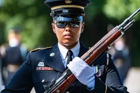 Soldier with sunglasses on carrying a rifle with bayonet affixed in front of her body at the diagonal is facing the camera.