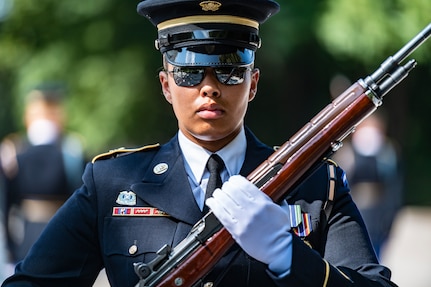 Soldier with sunglasses on carrying a rifle with bayonet affixed in front of her body at the diagonal is facing the camera.