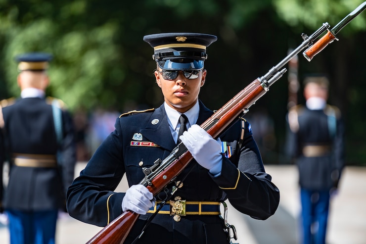 Woman in an Army dress uniform is carrying a rifle with bayonet affixed in front of her body diagonally while marching.