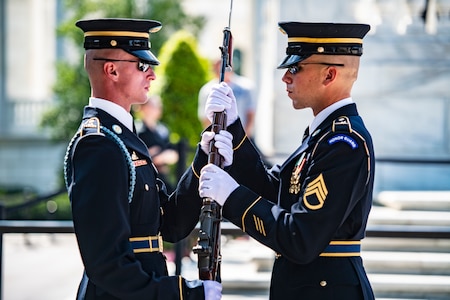 One soldier in Army dress uniforms is handing another soldier a rifle that has a bayonet affixed to it. They are performing a changing of the guard at the Tomb of the Unknown Soldier.